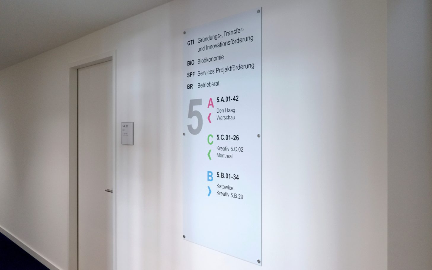Informationsschild mit Raumnummern im Flur eines Bürohaus.