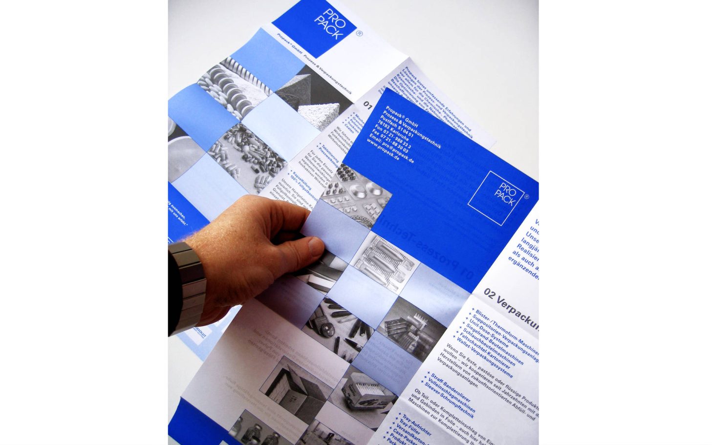 Eine Hand hält einen Flyer mit blauen quadratischen Flächen und Bildern.