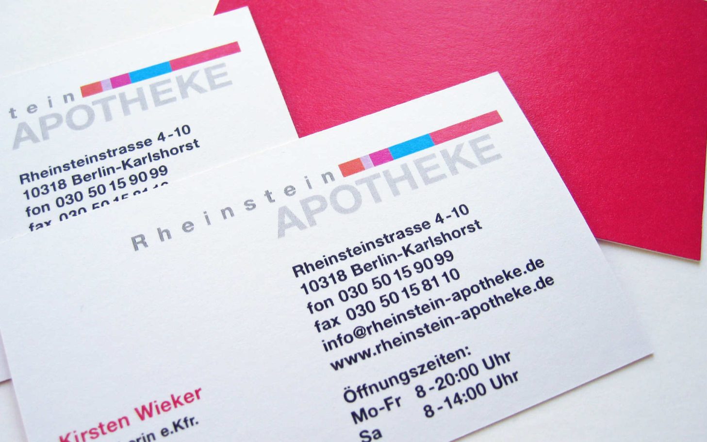 Zwei Visitenkarten der Rheinstein Apotheke mit mehrfarbigen Logobalken, Text und roter Rückseite.