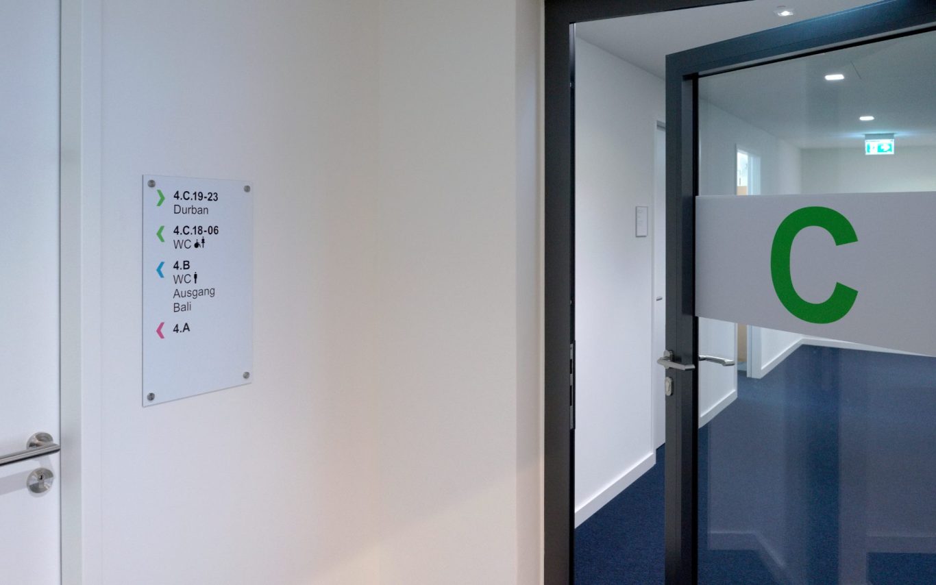 Flur-Schild mit Raumnummern und farbigen Pfeile. Auf Flurglastür großer grüner Buchstaben "C" 