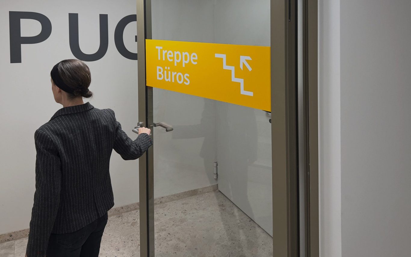 Gestaltung Richtungsbeschriftung und Piktogramme auf Glastüren Parkhauszugänge Bürohaus Schönefeld Eine Person öffnet eine Glastür mit einer gelben Richtungbeschriftung und Treppen-Symbol.